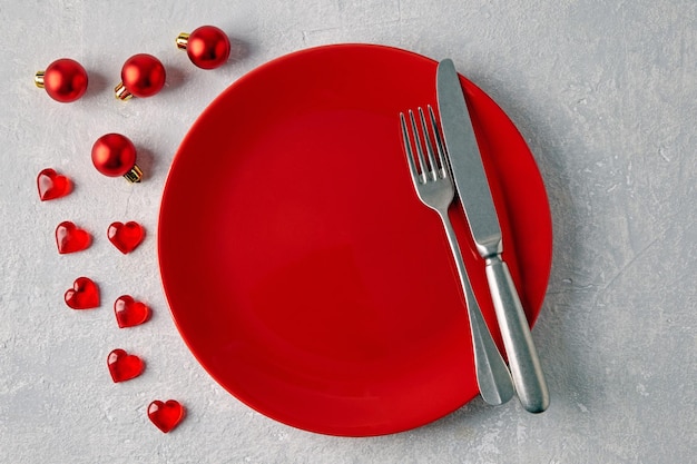 Un plato de cerámica rojo vacío con cubiertos rodeado de bolas de Navidad rojas y corazones en una mesa de hormigón claro