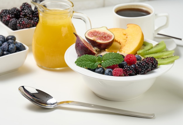 Plato completo con avena y frutas, jugo recién exprimido en una jarra de vidrio transparente, taza de café sobre una mesa blanca. Desayuno saludable
