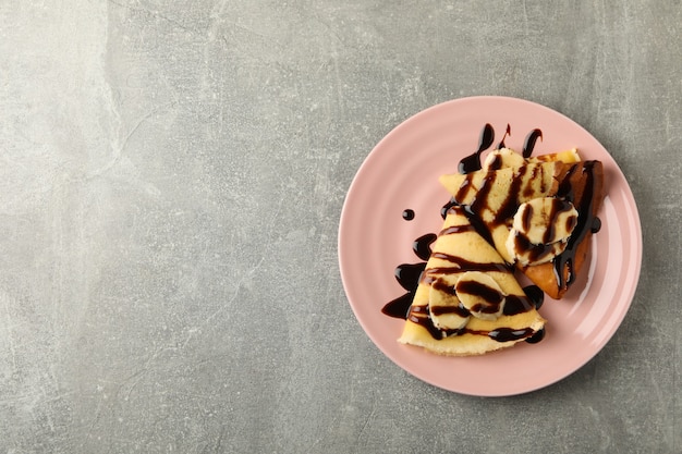 Plato de crepes con salsa de chocolate y rodajas de plátano sobre fondo gris