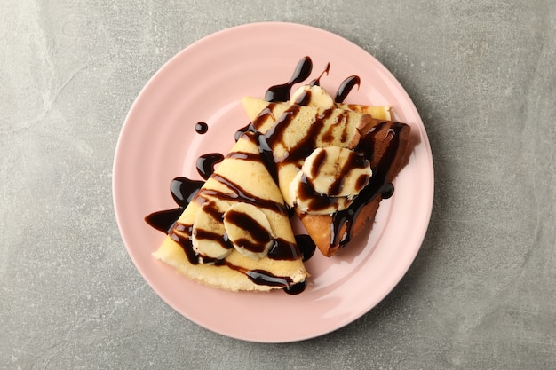 Plato de crepes con salsa de chocolate y rodajas de plátano sobre fondo gris