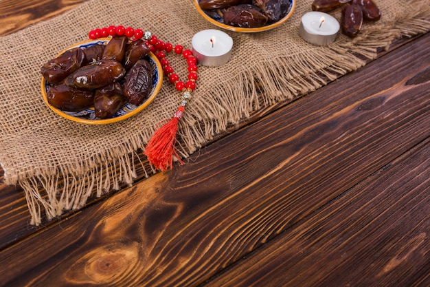 Plato de dátiles maduros con cuentas de oración rojas con velas encendidas sobre mantel de yute sobre el escritorio de madera