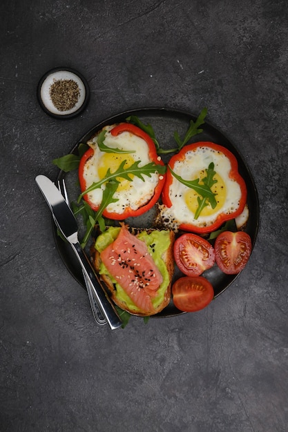 Plato de desayuno con huevos fritos sándwich de salmón con pimiento rojo y aguacate sobre fondo negro