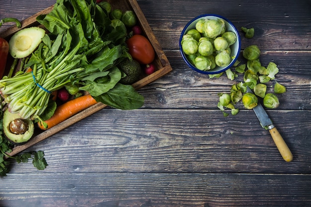 Foto un plato con diferentes verduras de temporada colocadas en la esquina de una mesa de madera y algunos dientes de bruselas a un lado pelados con un cuchillo