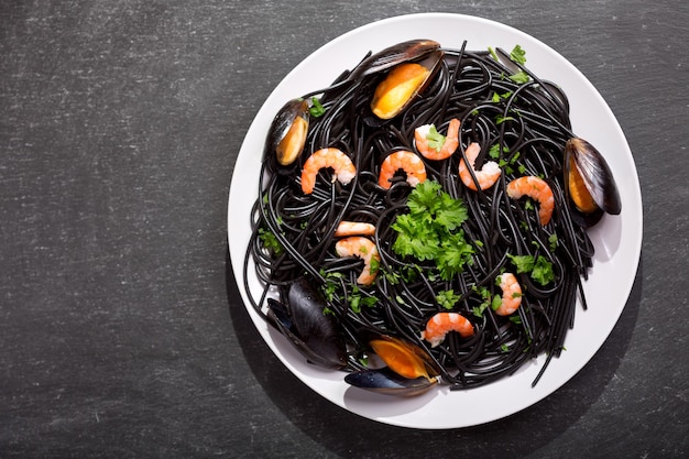 Plato de espaguetis negros con tinta de calamar con gambas, mejillones y perejil sobre mesa oscura, vista superior