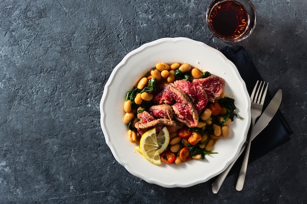 Plato con filete de res con frijoles blancos, espinacas, tomates y una copa de vino tinto.