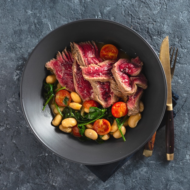 Plato con filete de ternera con frijoles blancos, espinacas y tomates sobre fondo de piedra oscura vista superior con co gratis