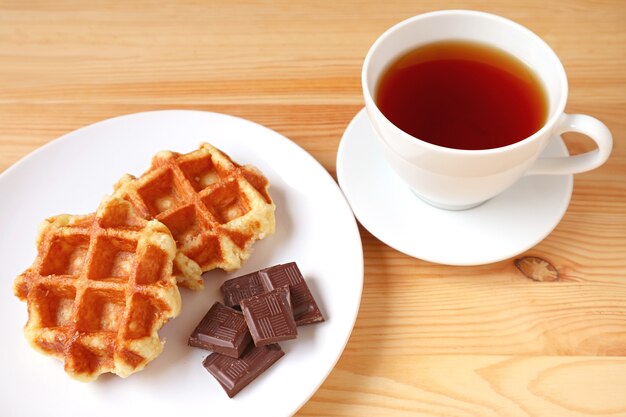 Plato de gofres de Lieja con trozos de chocolate negro y una taza de té caliente en la mesa de madera