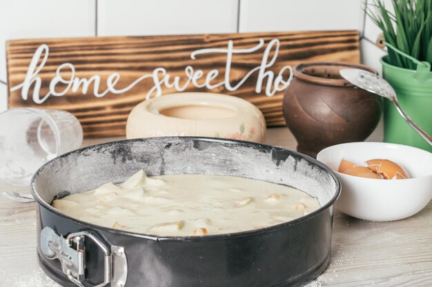 Plato para hornear con masa de tarta de manzana cruda se encuentra en la encimera de la cocina junto a los ingredientes, utensilios de cocina y letrero de madera Hogar, dulce hogar