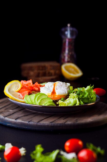 Plato con huevos pashot salmón aguacate y verduras Delicioso desayuno en tablero de madera oscura y fondo negro Variedad en comer para el buen humor