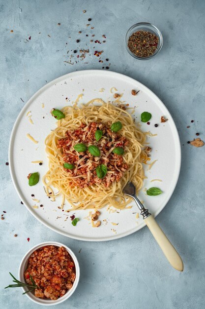 Plato italiano de pasta a la boloñesa en un plato plano, albahaca, especias y una servilleta ligera sobre un fondo gris.