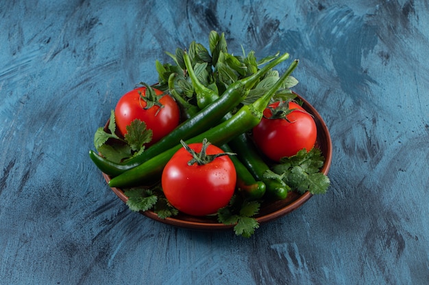 Plato de madera de tomates, chiles y verduras sobre superficie azul.