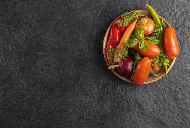 Plato de madera con verduras frescas pimiento verde tomate cebolla zanahoria pepino albahaca eneldo sobre fondo negro gris vista superior