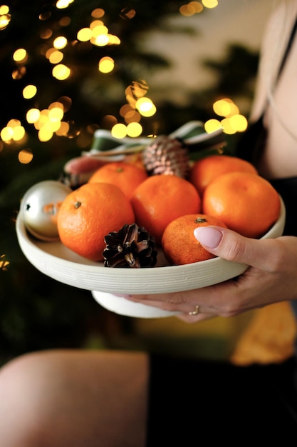 Plato con mandarinas y juguetes navideños en manos femeninas contra el fondo de las luces navideñas