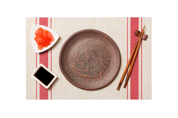 Plato marrón redondo vacío con palillos para sushi y salsa de soja, jengibre sobre fondo de estera de sushi. Vista superior con espacio de copia para su diseño.
