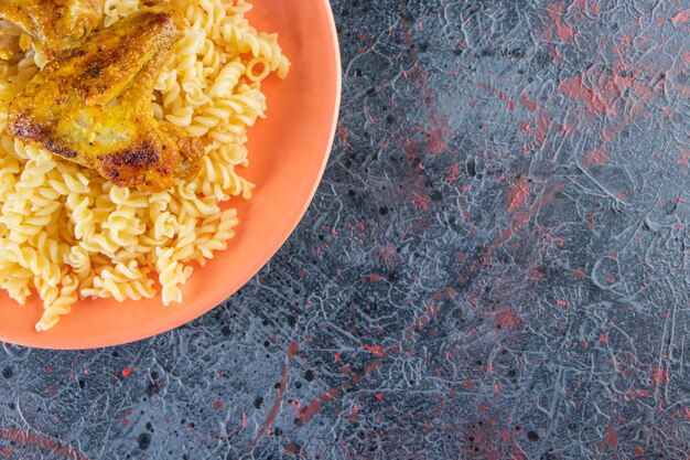 Plato naranja de pasta fusilli con alitas de pollo frito sobre superficie de mármol.