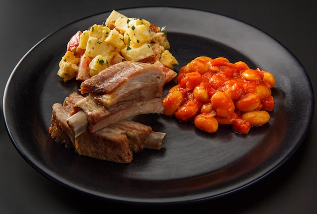 Plato negro con costillas al horno Frijoles en salsa y patatas aislado sobre un fondo negro