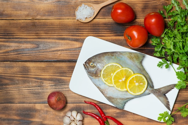 Plato de pescado cocinado con varios ingredientes. Pescado crudo fresco decorado con rodajas de limón y hierbas sobre mesa de madera.