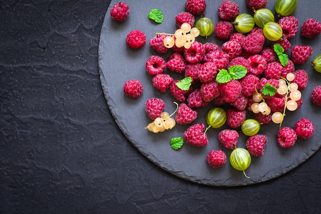 Plato de pizarra con diferentes frutos del bosque. Frambuesas, grosellas, grosellas sobre un fondo negro.