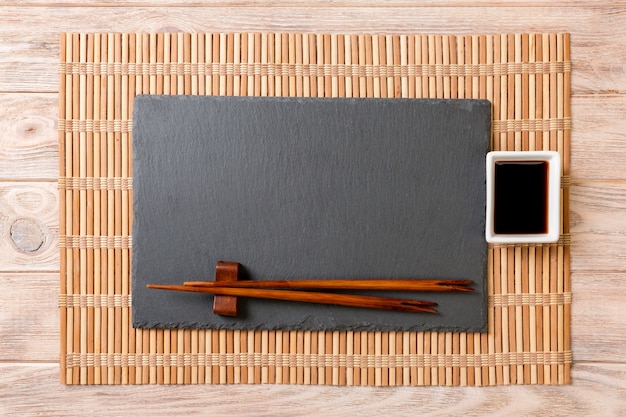 Plato de pizarra negro rectangular vacío con palillos para sushi y salsa de soja sobre madera