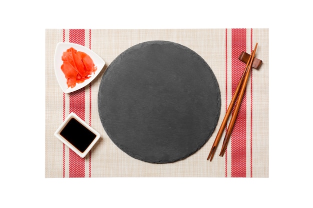 Plato de pizarra vacío redondo negro con palillos para sushi y salsa de soja, jengibre sobre fondo de estera de sushi. Vista superior con espacio de copia para su diseño.