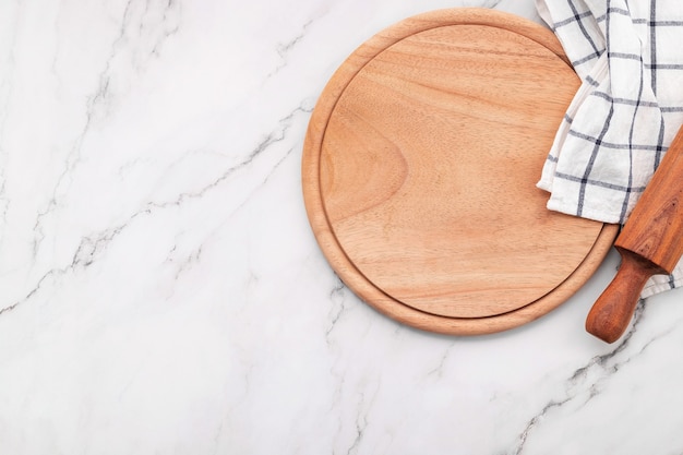 Plato de pizza de madera vacía con servilleta y rodillo en la mesa de la cocina de piedra de mármol. Tablero de pizza y mantel sobre fondo de mármol blanco.