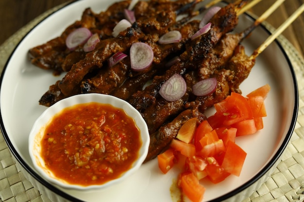 Un plato de pollo satay servido en un plato blanco con cebollas en rodajas, tomates y un bol sambal