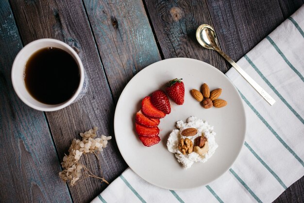 Plato con queso cottage, fresas y nueces, una taza de café y toallas en la mesa de madera, comida saludable, desayuno