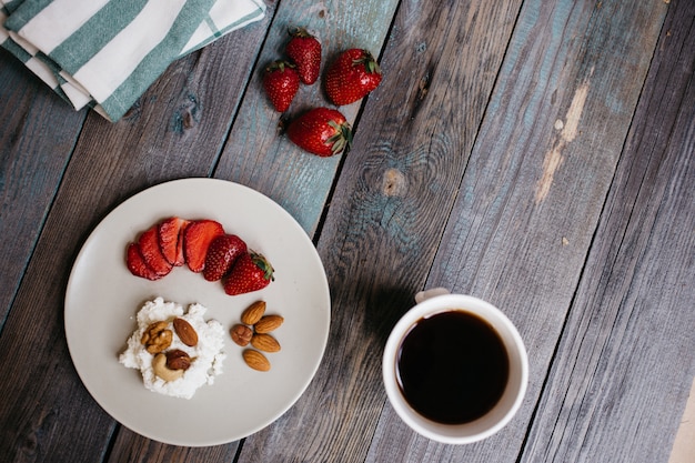 Plato con queso cottage, fresas y nueces, una taza de café y toallas en la mesa de madera, comida saludable, desayuno