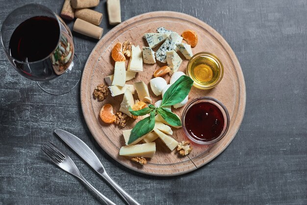 Plato de queso delicioso aperitivo para vino: queso, miel, nueces, mermelada, servido sobre una tabla de madera clara y un vaso con vino tinto sobre una superficie de madera