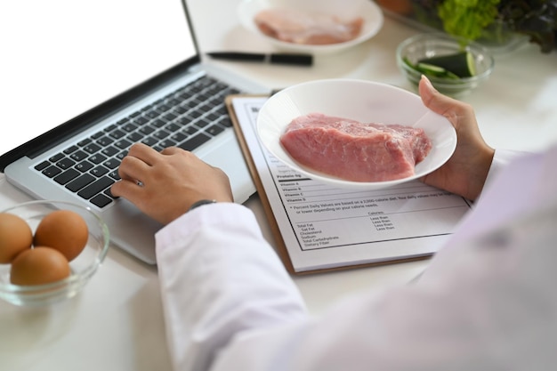 Plato de retención de nutricionista con carne cruda dando consulta en línea al paciente a través de una computadora portátil