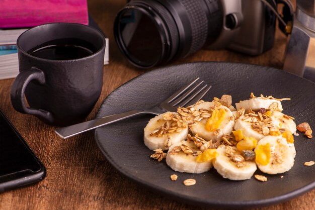 Plato con rodajas de plátano, granola, fruta confitada y miel, tostadas y una taza de café.