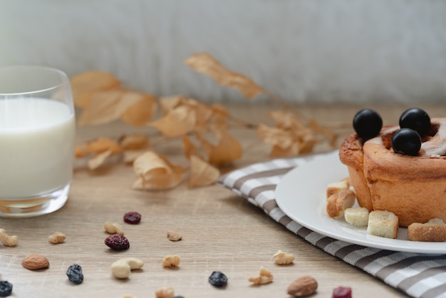 Plato con sabroso bollo de canela al horno con uvas negras en la parte superior, un vaso de leche y muchas nueces mixtas con pasas en la mesa de madera