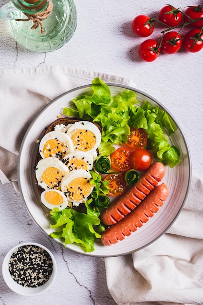 Foto plato con salchichas, sándwiches de huevo, tomates y hojas de lechuga en la mesa y vista vertical