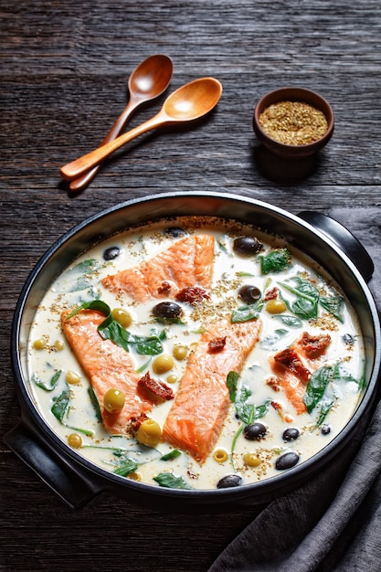 Plato de salmón toscano de filete de salmón con piel a la sartén con salsa de crema, ajo, puerro, tomates secados al sol, aceitunas y espinacas en una fuente para hornear negra sobre una mesa de madera oscura, vista superior, primer plano