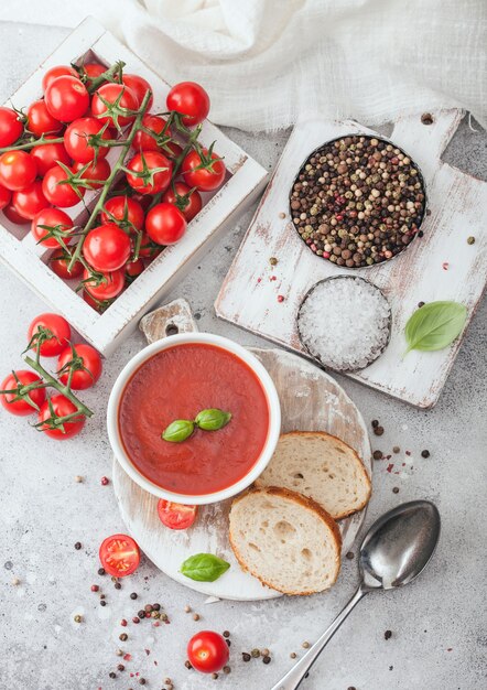 Plato de tazón de fuente blanco de sopa de tomate cremosa con cuchara sobre mesa de luz con caja de tomates crudos y pan. Vista superior