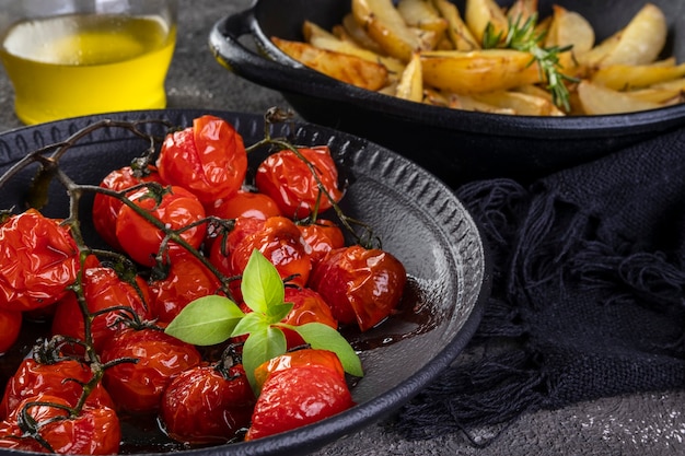 Plato de tomates confitados y patatas asadas al romero en cazuela de hierro sobre fondo oscuro.