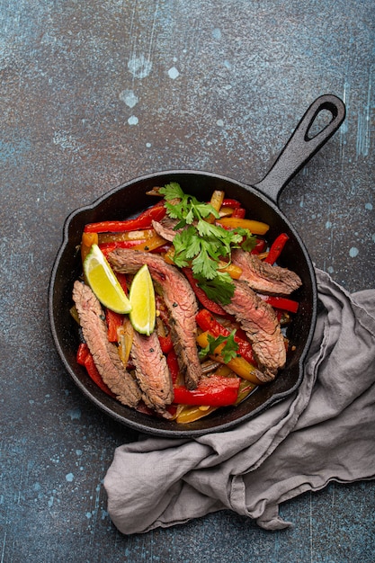 Plato tradicional mexicano fajitas de carne con pimientos en sartén de hierro fundido negro sobre fondo de piedra rústica desde arriba, comida mexicana americana