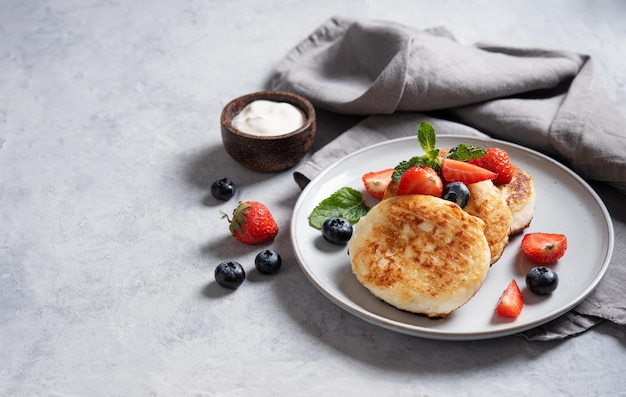 Plato tradicional ruso para el desayuno syrniki. Tortitas de requesón con crema agria y bayas frescas sobre un fondo azul con una servilleta. Orientación horizontal.