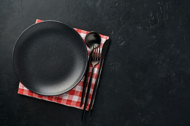 Plato vacío negro, tenedor y cuchillo y mantel rojo. Utensilios de cocina en mesa de piedra negra.