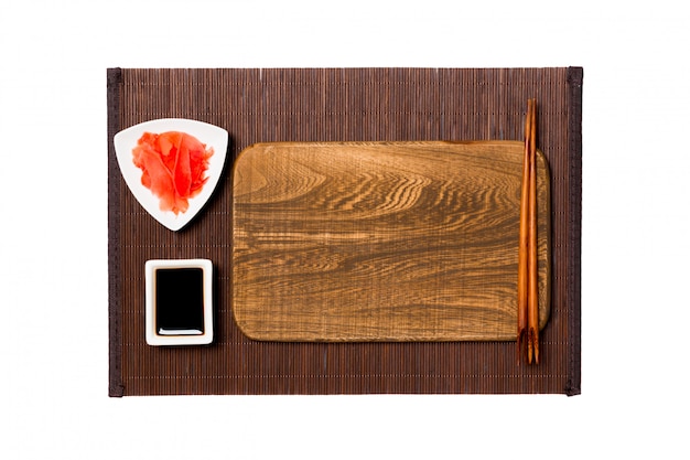 Plato vacío rectangular de madera marrón con palillos para sushi, jengibre y salsa de soja sobre estera de bambú oscuro. Vista superior con copyspace