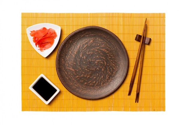 Plato vacío redondo de color marrón con palillos para sushi, jengibre y salsa de soja sobre estera de bambú amarillo