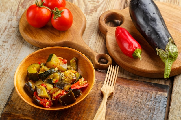 Un plato de verduras asadas junto a tomates y berenjenas sobre tablas de madera junto a un tenedor de madera