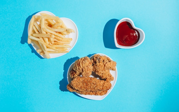 Platos en forma de corazón con bocadillos sobre un fondo azul nuggets alas tiras y papas fritas con ketchup sol sombras duras