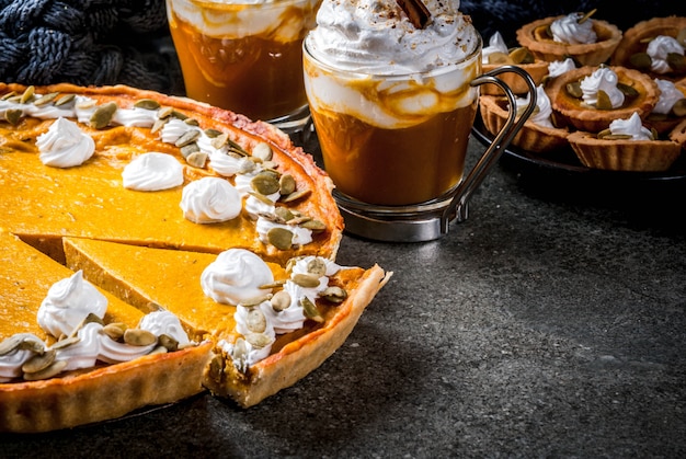 Platos tradicionales de otoño. Halloween, Acción de Gracias. Pastel de calabaza, tartaletas de calabaza con crema batida y semillas de calabaza, café con leche de calabaza con canela en una mesa de piedra negra con una manta. Copia espacio