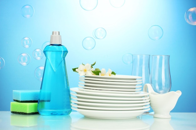 Platos y vasos limpios y vacíos con esponjas líquidas para lavar platos y flores sobre fondo azul.