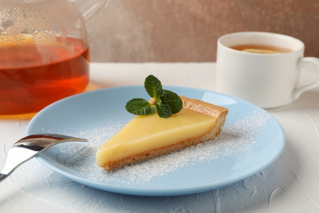 Platte mit Zitronentörtchenscheibe, Teekanne und Tasse Tee auf weißem Tisch, Nahaufnahme