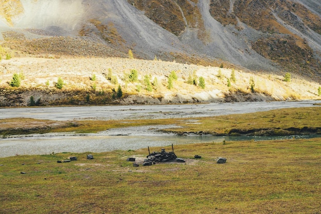 Platz für Lagerfeuer in der Nähe des Bergflusses mit Blick auf den sonnenbeschienenen Berghang. Campingplatz in der Nähe von Fluss und Nadelbäumen am Hang bei goldenem Sonnenschein im Herbst. Bunte Berglandschaft in strahlender Sonne.