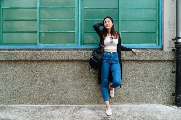 Platz kopieren. Ganzaufnahme der schönen lächelnden asiatischen Frau, die sich an Wand und hölzernes Fensterbrett im Freien lehnt. charmante dame mit tasche schnippt haare und schaut zur seite und genießt das sonnenlicht