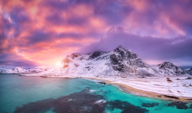 Playa de arena nórdica con mar azul en invierno al atardecer en las islas Lofoten Noruega Paisaje con montañas nevadas cielo violeta con nubes rosas pueblo de agua con edificios gente caminando Naturaleza