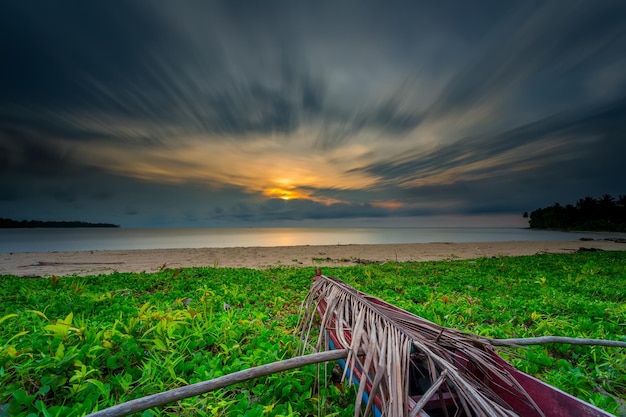 Una playa con un barco en primer plano y una puesta de sol al fondo.